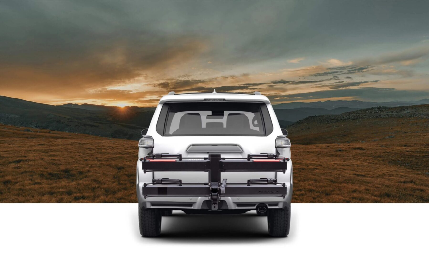 A white SUV with a rear-mounted rack is parked on a grassy plain, facing a scenic sunset with mountains in the background.