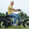 A man rides the Meet One Tour Dual Motor Dual Battery Electric Trike on grass, dressed in a yellow polo, blue jeans, and gray sneakers. Trees and a cloudy sky are visible in the background.