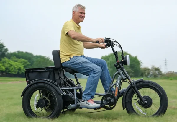 A man rides the Meet One Tour Dual Motor Dual Battery Electric Trike on grass, dressed in a yellow polo, blue jeans, and gray sneakers. Trees and a cloudy sky are visible in the background.