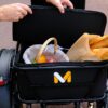 A person lifts the lid of a black cargo basket on the Meet One Tour Dual Motor Dual Battery Electric Trike, revealing a wicker basket filled with fruit and two baguettes in a paper bag.
