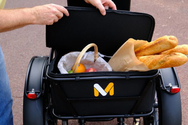 A person lifts the lid of a black cargo basket on the Meet One Tour Dual Motor Dual Battery Electric Trike, revealing a wicker basket filled with fruit and two baguettes in a paper bag.