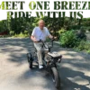 An older man rides a Meet One Breeze Folding Electric Trike on a paved driveway surrounded by greenery. Large text above reads, “MEET ONE BREEZE WITH ME.”.