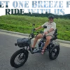 A man in sandals and sunglasses sits on a Meet One Breeze Pro Fat Tire Electric Trike on a paved road, surrounded by greenery and hills. Text above reads “MEET ONE BREEZI PRO - RIDE WITH US.”.