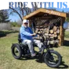 An older man in a blue hoodie and cap sits on a Meet One Tour Lite Full Suspension Electric Trike in front of a woodshed labeled "She Shed" and "He Sheds" on a sunny day.