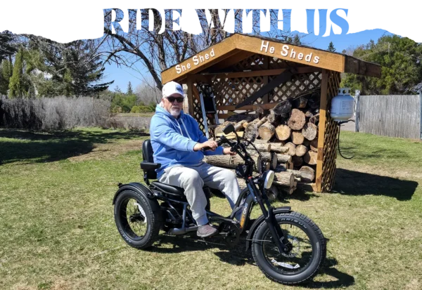 An older man in a blue hoodie and cap sits on a Meet One Tour Lite Full Suspension Electric Trike in front of a woodshed labeled "She Shed" and "He Sheds" on a sunny day.