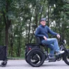 A man in a helmet sits on a Meet One Tour Dual Motor Dual Battery Electric Trike with a black cargo trailer, parked on a paved path lined with trees.