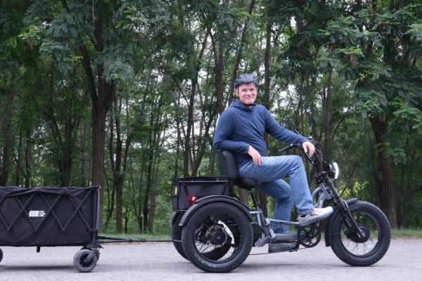 A man in a helmet sits on a Meet One Tour Dual Motor Dual Battery Electric Trike with a black cargo trailer, parked on a paved path lined with trees.