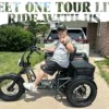 A man in sunglasses and a gray tank top sits on the Meet One Tour Lite Full Suspension Electric Trike in a driveway, flashing a peace sign. A white SUV and greenery are visible in the background.