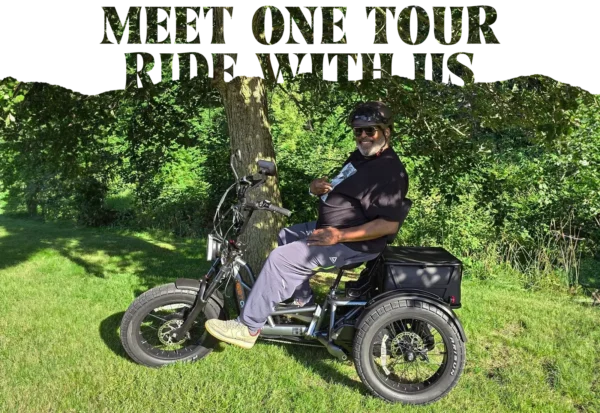 A man wearing a helmet sits on a Meet One Tour Dual Motor Dual Battery Electric Trike under a tree in a grassy park.