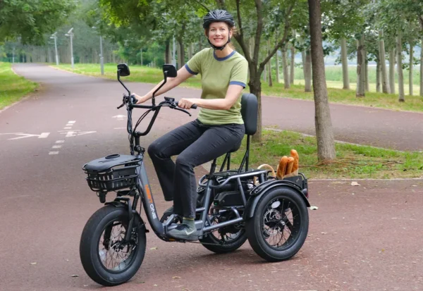 A person wearing a helmet sits on a Meet One Breeze Folding Electric Trike on a paved park path, with a basket holding two baguettes behind the seat.
