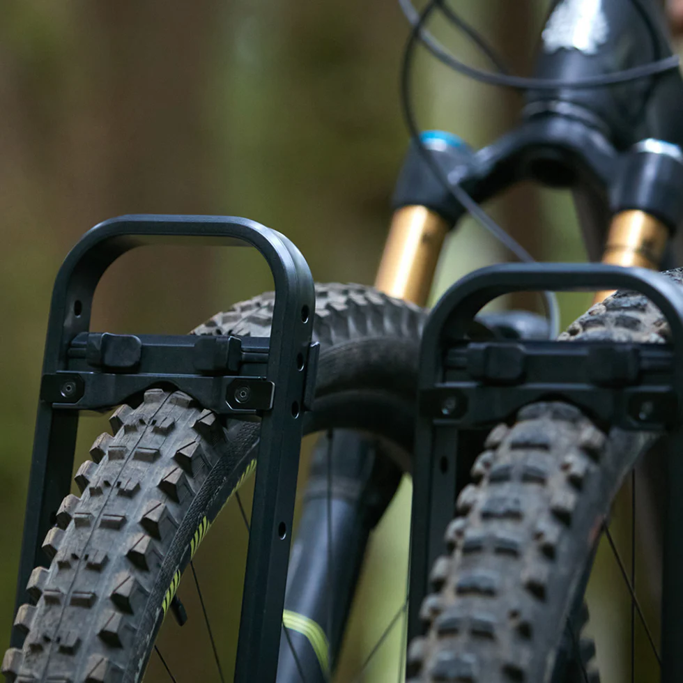 Close-up of two mountain bike tires with black frame racks attached, set against a blurred outdoor background.