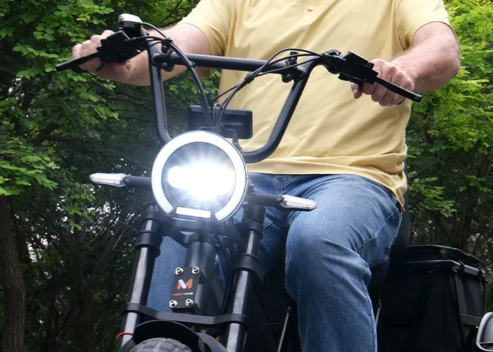 A person wearing a yellow shirt and jeans rides an electric bike with the headlight turned on, outdoors with trees in the background.