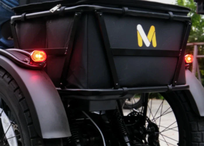 Close-up of the rear side of a three-wheeled vehicle with a black cargo box, red brake lights on, and a yellow "M" logo on the box.