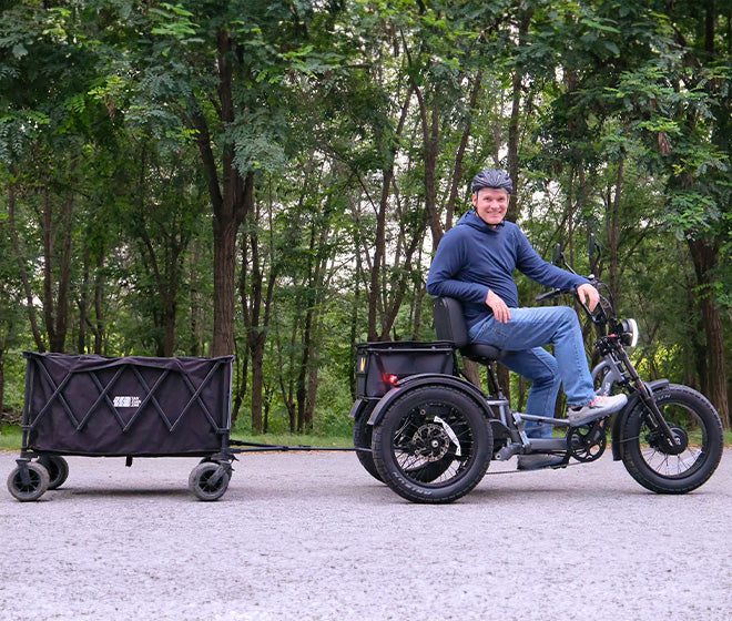 A person wearing a helmet rides an electric tricycle with a black cargo trailer attached, on a paved path surrounded by trees.