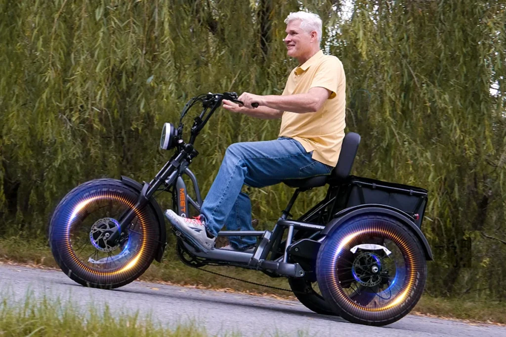 An older man rides an electric tricycle with large, illuminated front wheels along an outdoor path lined with trees.