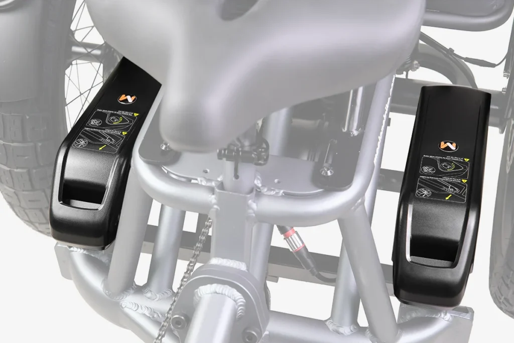 Close-up view of two black wheelchair battery packs mounted on either side of a metal frame beneath a wheelchair seat.