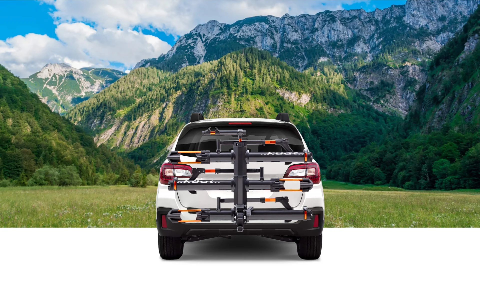A white SUV with a bike rack mounted on the rear is parked in front of a scenic mountain landscape with green trees and a blue sky.