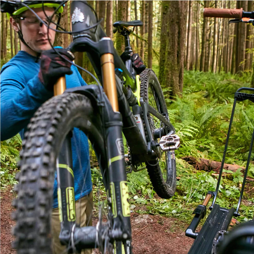 A person wearing a blue shirt and helmet lifts a mountain bike on a forest trail surrounded by trees and ferns.