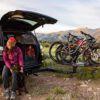 A person in outdoor gear sits by an SUV with the trunk open, petting a dog. Three mountain bikes are mounted on a Pivot™ v2 rear bike rack, with mountains visible in the background.