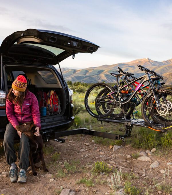 A person in outdoor gear sits by an SUV with the trunk open, petting a dog. Three mountain bikes are mounted on a Pivot™ v2 rear bike rack, with mountains visible in the background.