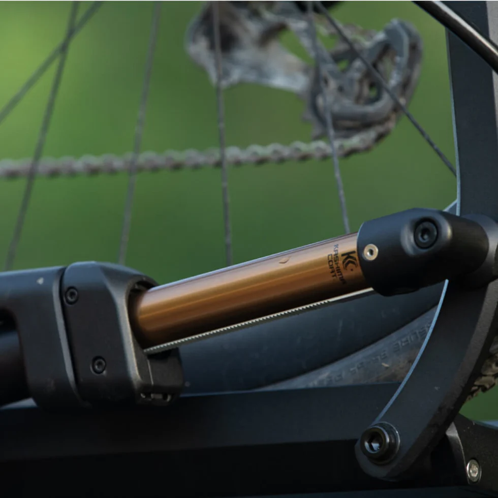 Close-up of a bicycle rear suspension system, showing the shock absorber with a gold-colored shaft and part of the rear wheel with visible chain and spokes.