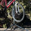 A red bicycle is mounted on the Tri Doc - Trail Doc Stand outdoors, highlighting the front wheel and fork, with trees and dirt in the background.