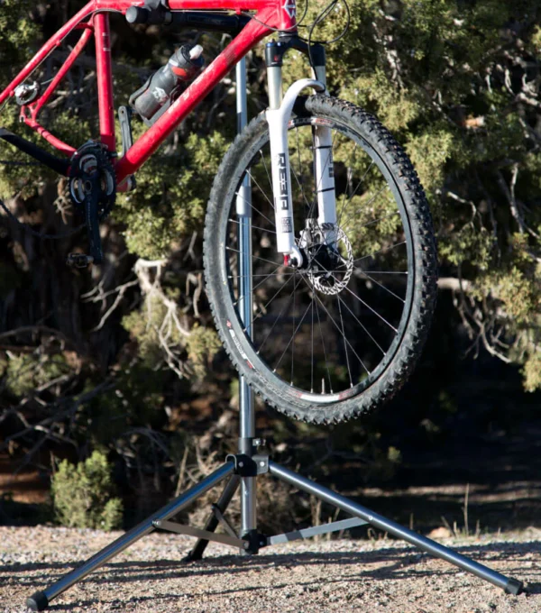 A red bicycle is mounted on the Tri Doc - Trail Doc Stand outdoors, highlighting the front wheel and fork, with trees and dirt in the background.