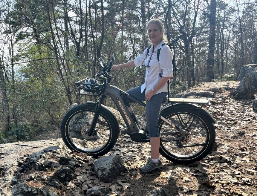 A woman wearing a white shirt and backpack stands beside an electric bicycle on a rocky forest trail under sunlight.