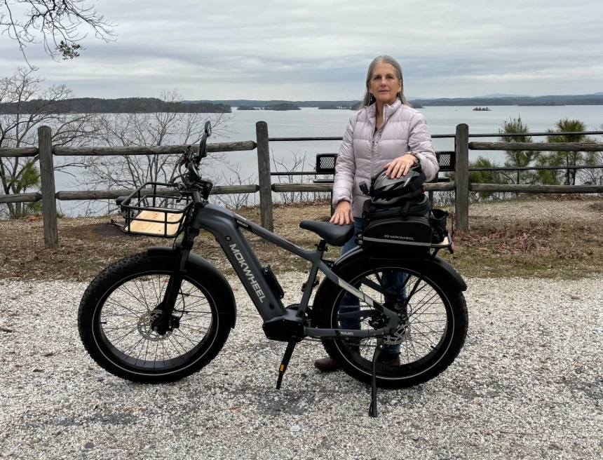 A woman stands beside a black e-bike with cargo racks, overlooking a lake and trees under a cloudy sky.