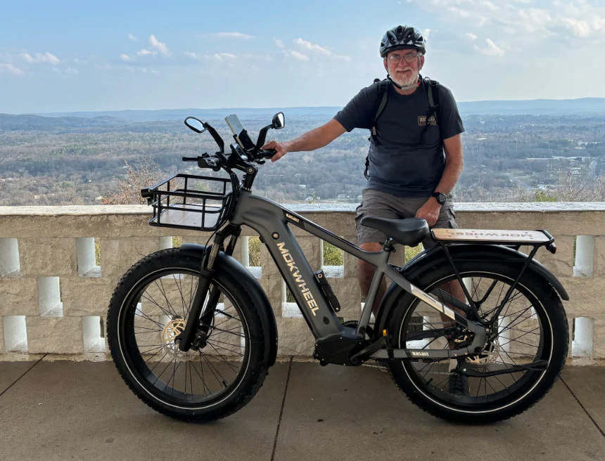An older man wearing a helmet and outdoor gear sits on a black electric bike labeled "ADDWHEEL" at a scenic overlook with a view of trees and hills.