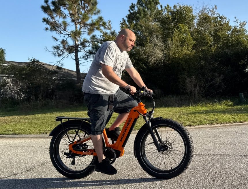 A man rides an orange electric bicycle on a suburban street with trees and houses in the background.