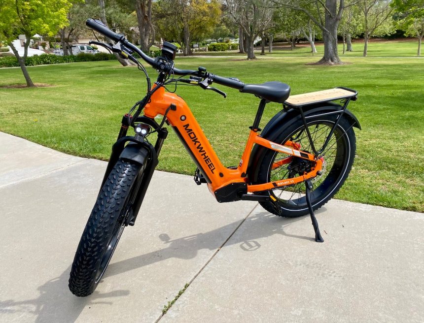 An orange Mokwheel electric bike with wide tires is parked on a concrete path in a park with grass and trees in the background.