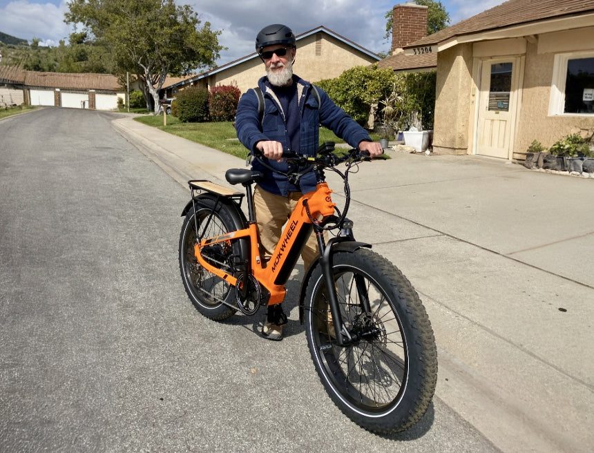 An older man wearing a helmet stands on a suburban street with an orange electric bicycle on a cloudy day.