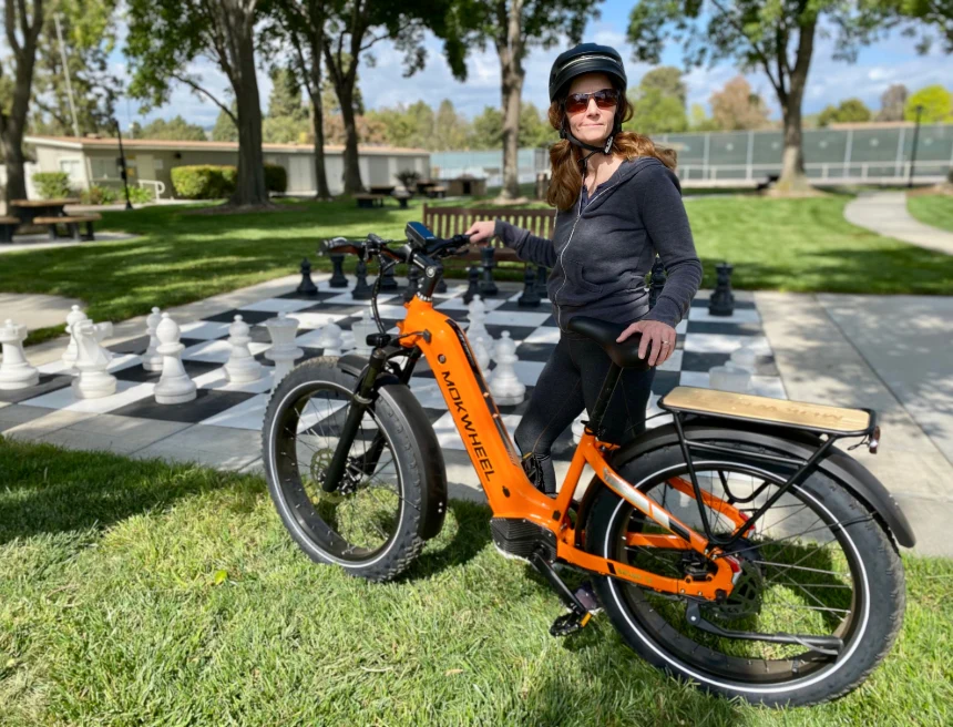 A person wearing a helmet sits on an orange electric bike in a park, near a large outdoor chessboard with oversized chess pieces.
