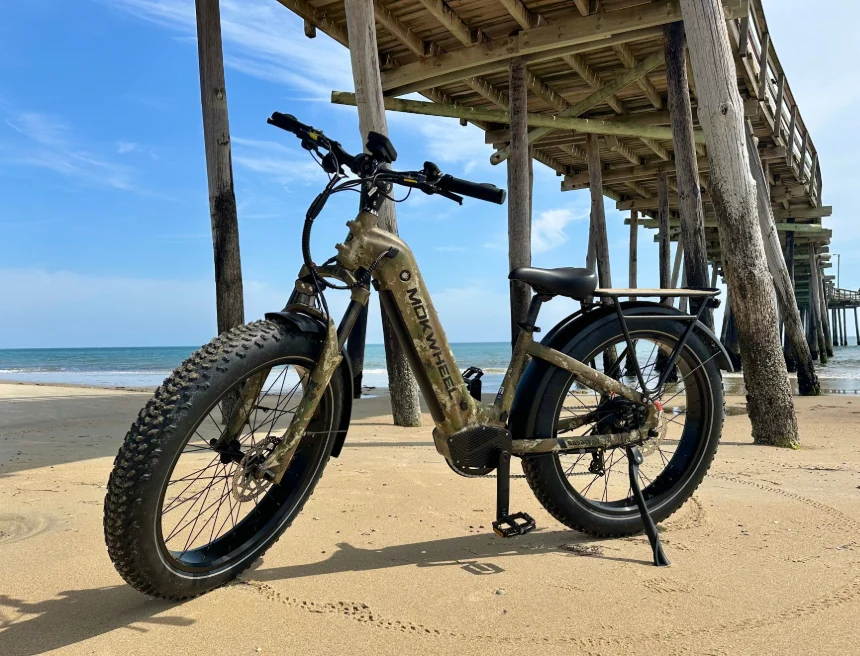 A fat-tire electric bicycle stands on sandy beach near the ocean, beside a wooden pier under a clear blue sky.