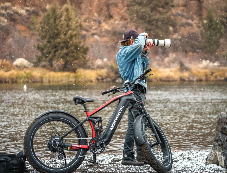 A person stands by a river taking photos with a camera and large lens, next to a red and black Mokwheel electric bike.