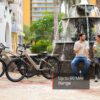 Two people relax with drinks by a fountain next to three parked Asphalt Ebikes. Text on the image reads, "Up to 60 Mile Range.