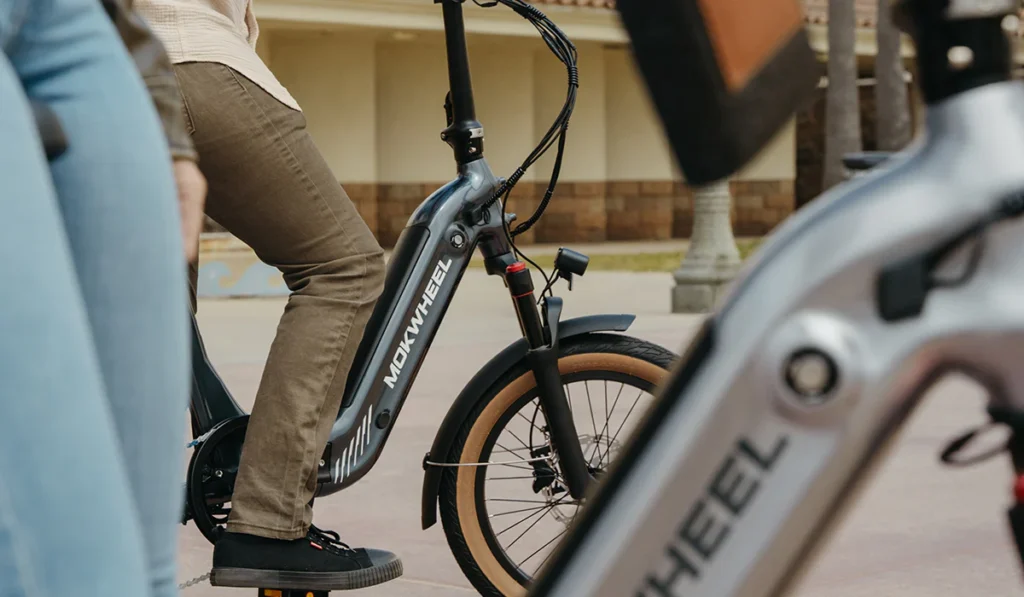 Two people on Mokwheel electric bikes are stopped outdoors, with focus on the bike frames and wheels. Only their lower bodies and the bikes are visible.