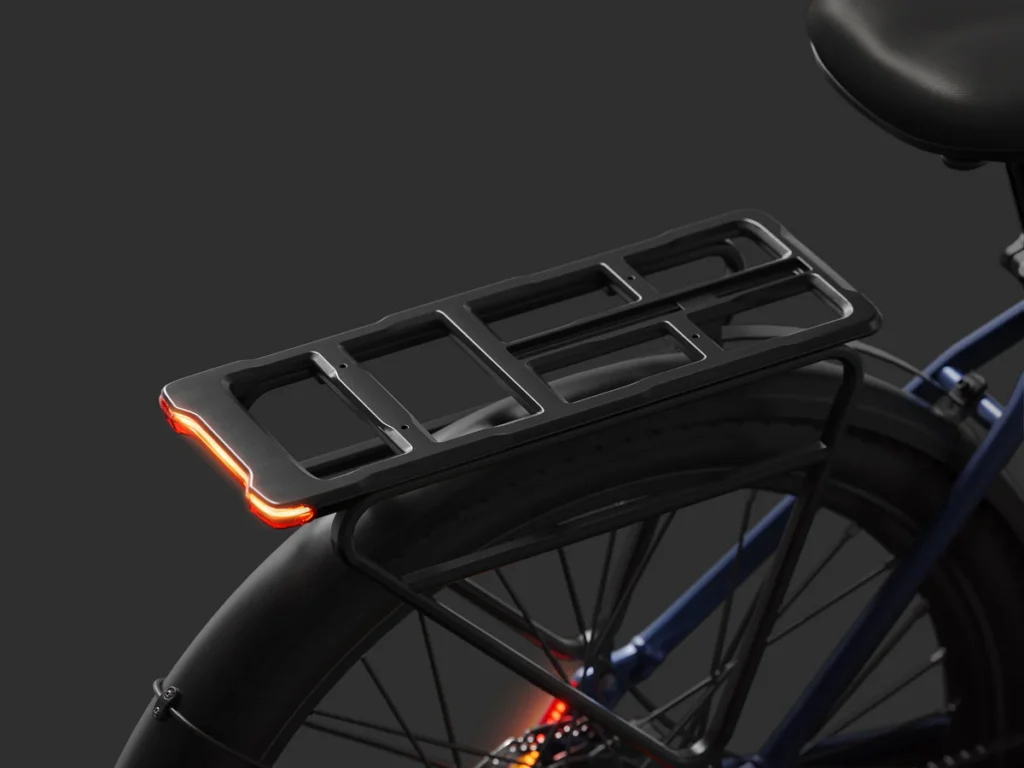 Rear view of a bicycle showing a black cargo rack with integrated red LED tail lights, attached above the back wheel on a dark background.