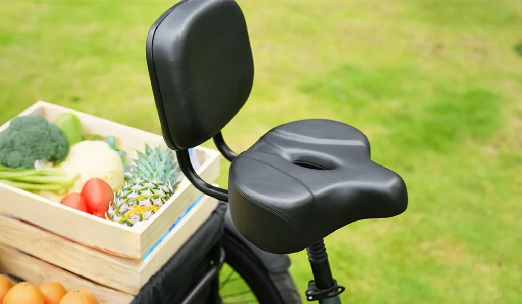 A black bicycle seat with a backrest is attached to a bike next to a wooden crate of vegetables and fruit, set on grass.