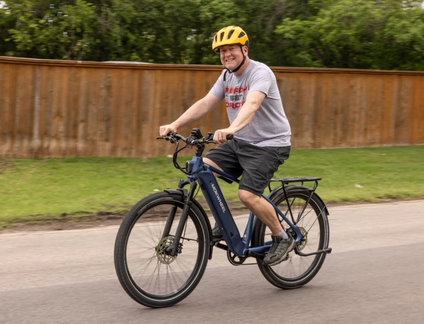 A man wearing a yellow helmet rides a blue electric bicycle on a paved road next to a wooden fence and grassy area.