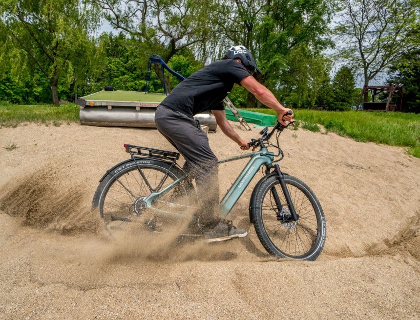 A person rides an electric bike through loose sand, causing the rear wheel to spin and kick up dust, in an outdoor park setting.