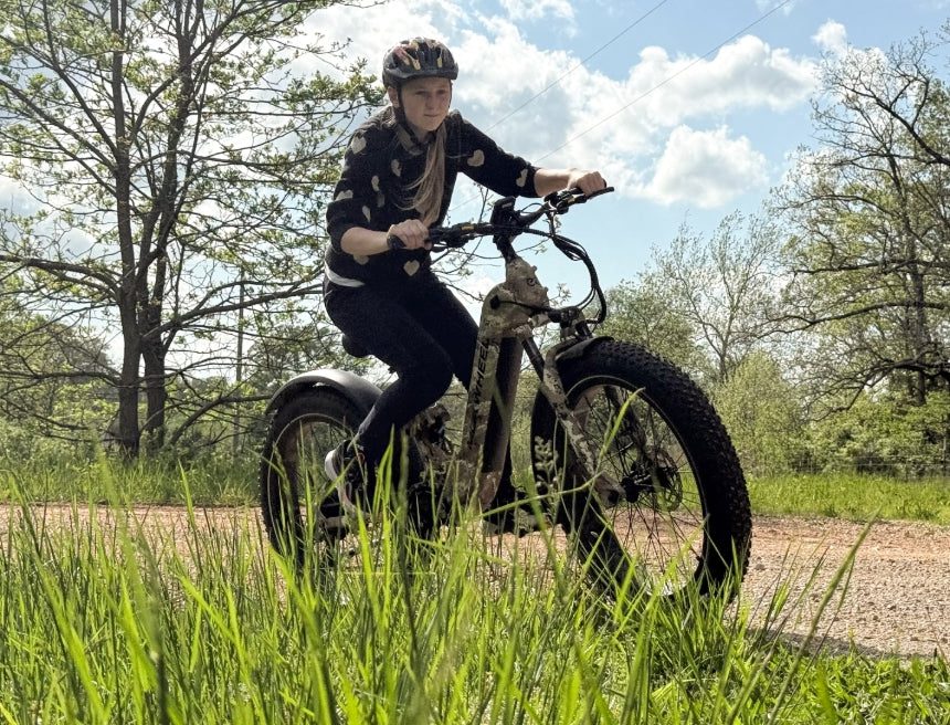 Person wearing a helmet rides a fat tire electric bike on a gravel path surrounded by greenery and trees under a partly cloudy sky.