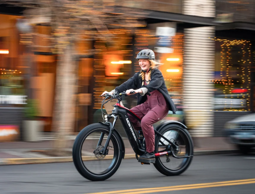 A person wearing a helmet and casual clothes rides an electric bicycle down a city street, with shops and lights blurred in the background.