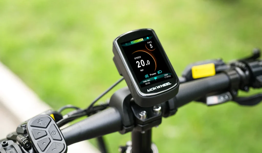 A close-up of a Mokwheel digital display mounted on a bicycle handlebar shows speed, battery level, and gear information. Grass is visible in the blurred background.