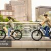 Two people wearing helmets ride Tarmac ST Ebikes across a city bridge with buildings and trees behind them. Text overlay reads "Up to 60 Mile Range.