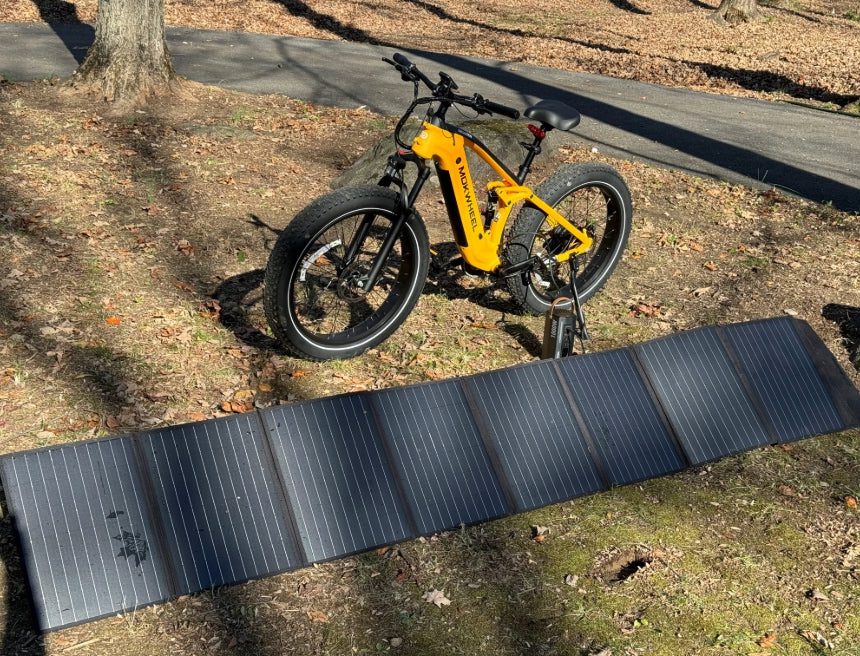 A yellow electric bicycle is parked on grass next to a large, unfolded solar panel under sunlight in a wooded area.