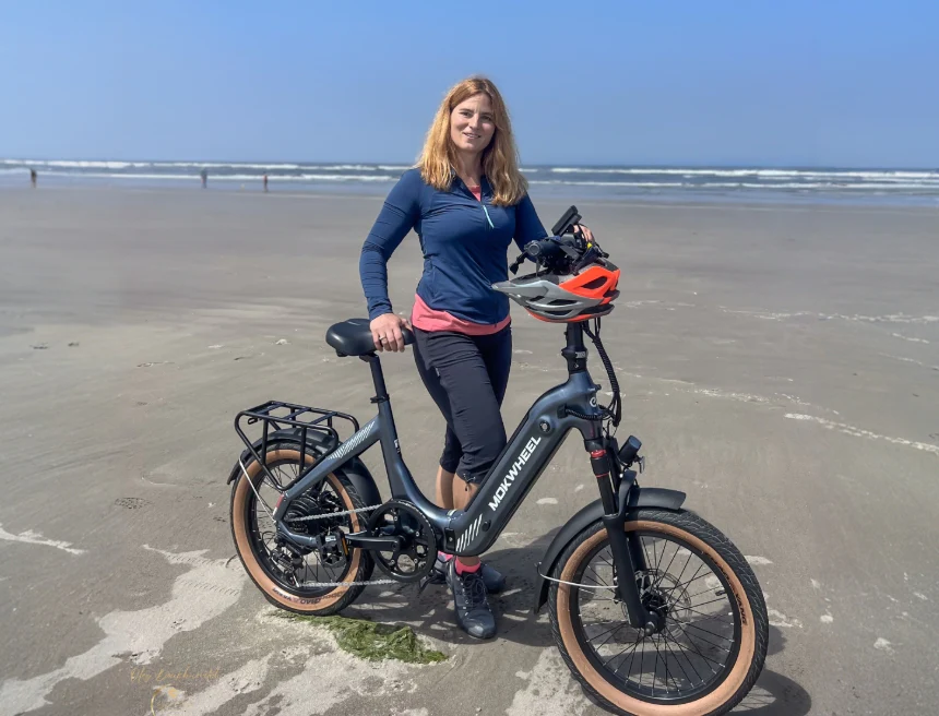 A person stands on a sandy beach next to an electric bicycle, holding a helmet, with waves and blue sky in the background.
