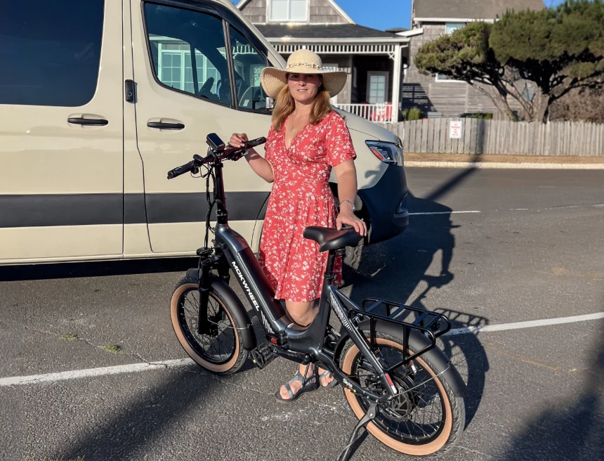 A woman in a red floral dress and hat stands next to a black electric bicycle in a sunny parking lot beside a beige van.