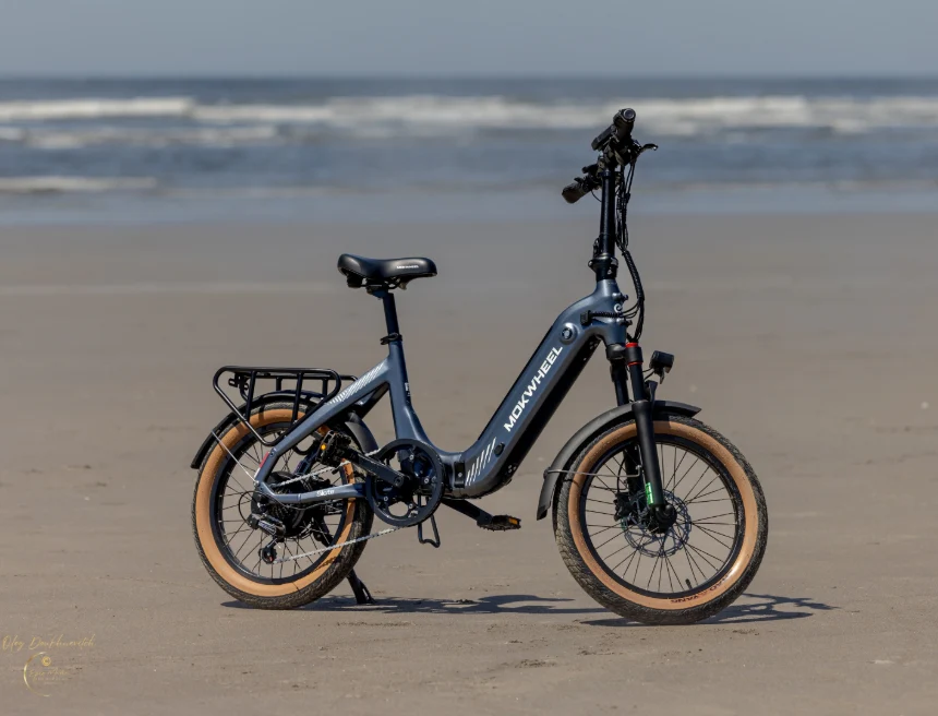 A blue folding electric bicycle with tan tires is parked on wet sand near the shoreline, with the ocean and sky in the background.